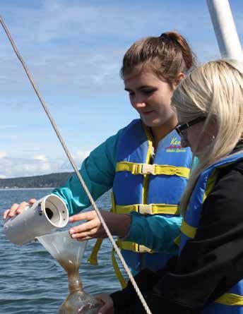 One student holds a cylinder cup in a pouring position. Another with long blonde hair holds a flask-like container beneath the cup. Both are on a boat wearing life jackets.